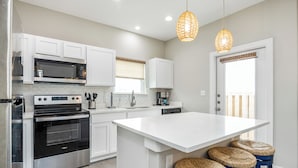 A modern kitchen with white cabinetry, stainless steel appliances, a large island with stools, and a hanging light fixture. - Coastal Family Escape | Shuffleboard, Heated Pools & Beach Nearby (Corpus Christi)