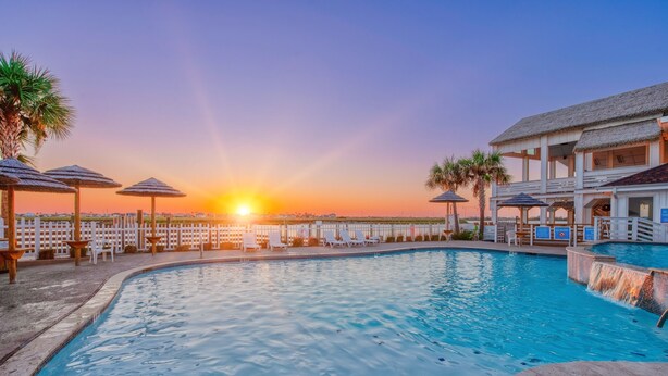 A swimming pool with lounge chairs and umbrellas, a building with a thatched roof, and a sunset in the background. - Coastal Family Escape | Shuffleboard, Heated Pools & Beach Nearby (Corpus Christi)