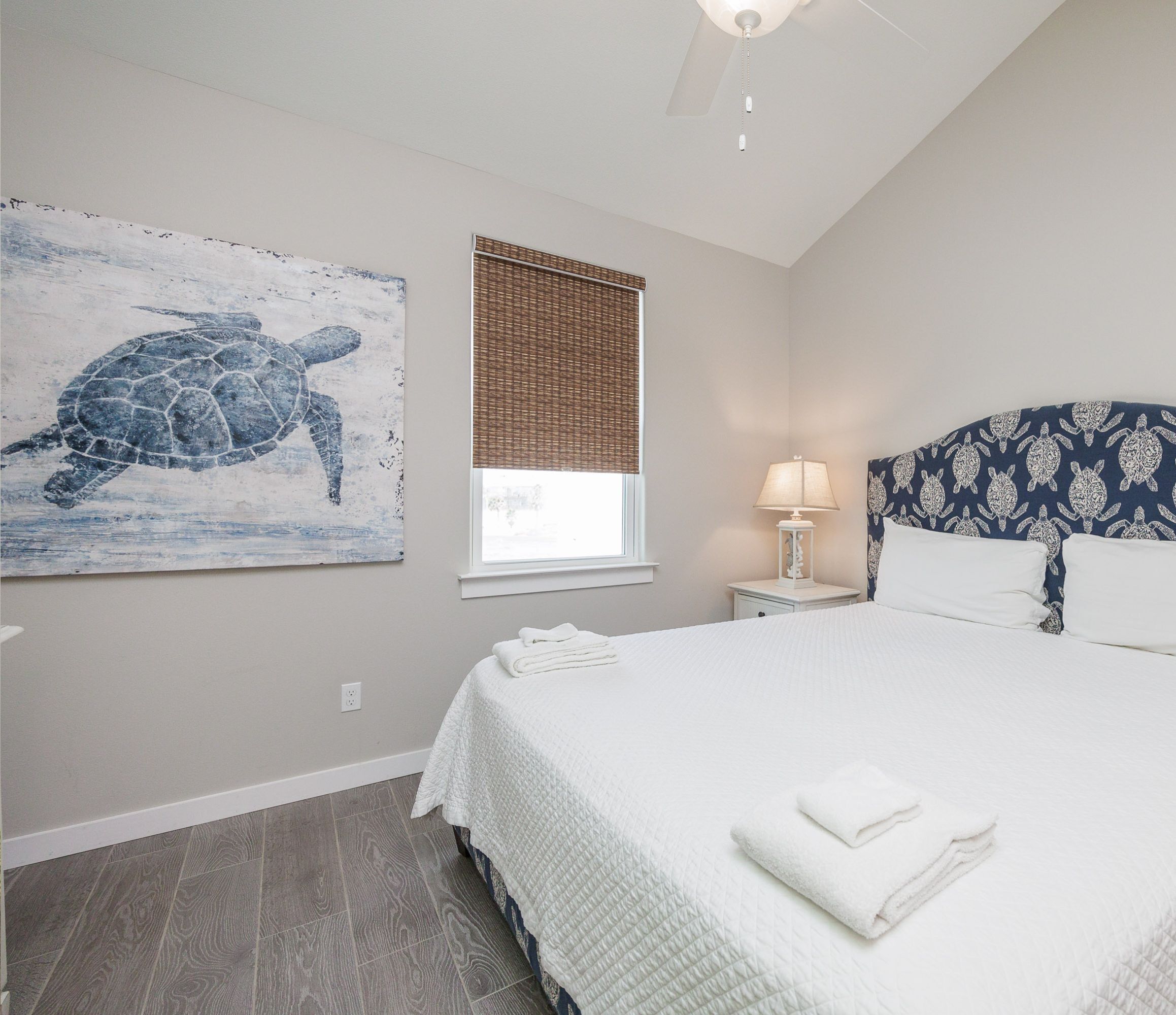 A bedroom with a large bed, a wall-mounted artwork of a turtle, a window with blinds, a bedside lamp, and a headboard with a turtle pattern.