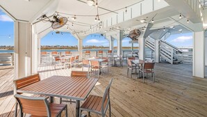 A covered outdoor dining area with wooden tables and chairs, string lights, and a view of the sea. - Pet-Friendly Coastal Stay | Pools & Waterfront Access (Corpus Christi)