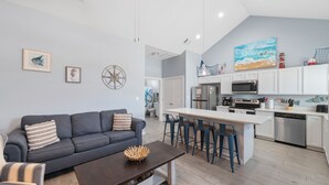 A modern kitchen with white cabinets, a central island with bar stools, a dark-colored sofa with striped pillows, and a dining area with a wooden table. - Pet-Friendly Coastal Stay | Pools & Waterfront Access (Corpus Christi)
