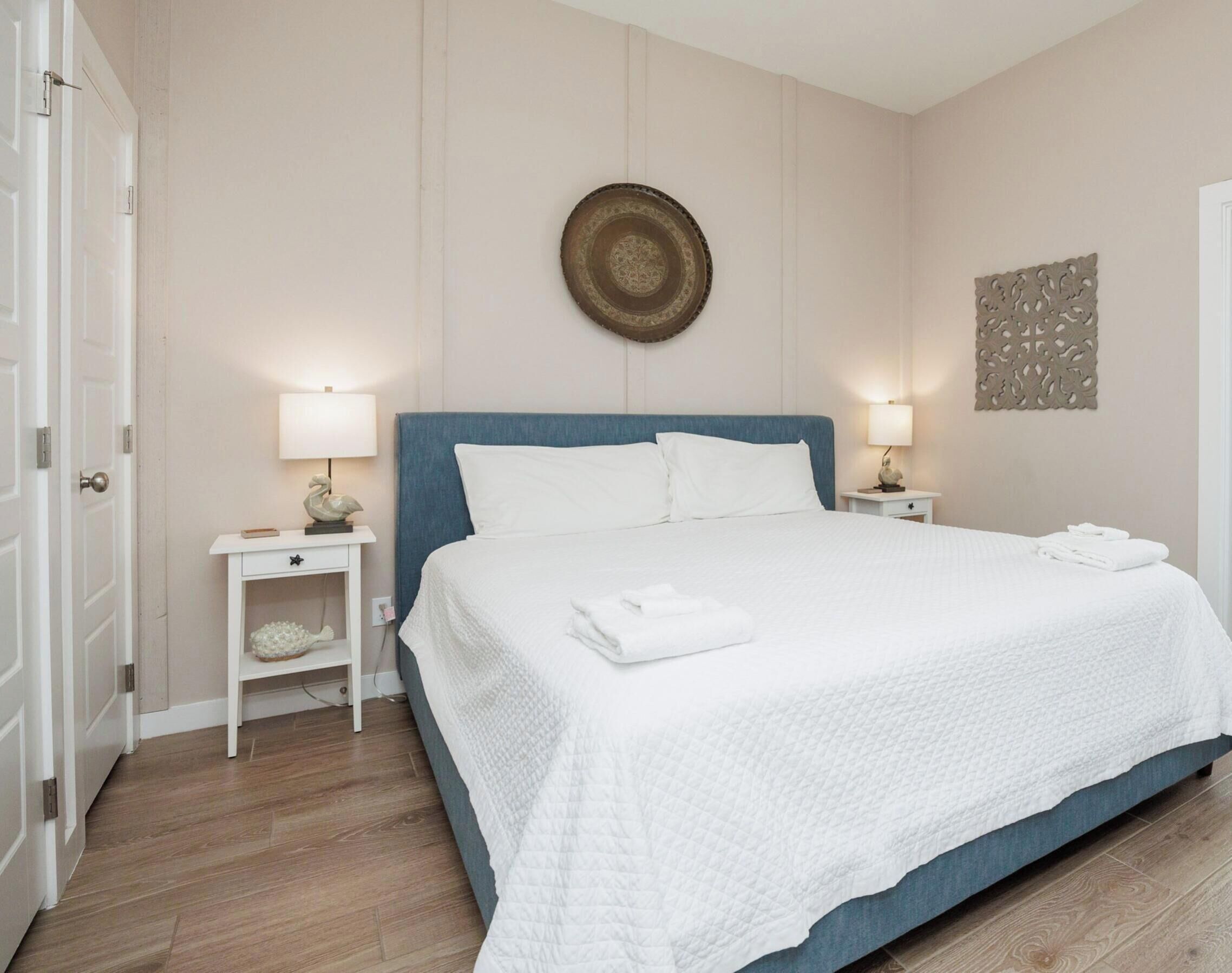 A neatly made bed with white linens, two bedside tables with lamps, a decorative wall piece, and a wooden floor.
