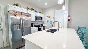 A modern kitchen with a stainless steel refrigerator, microwave, and stove. There is a sink with a faucet, and a teal chair is visible. - Family Beach Getaway | 2 Pools, Splash Pad & Pets Welcome (Corpus Christi)