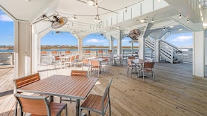 A covered outdoor dining area with wooden tables and chairs, string lights, and a view of the sea.
