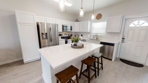 A modern kitchen with white cabinetry, stainless steel appliances, a central island with stools, pendant lighting, and a clock on the wall.