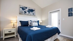 A neatly made bed with a blue bedspread and white linens, a white nightstand with a lamp, and a world map artwork above the bed. - Serene Family Townhome | Water Views, Pools, Docks & Pets Welcome (Corpus christi)