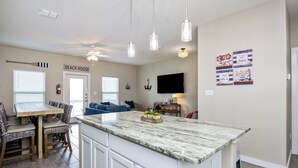 A modern kitchen with a large island, white cabinetry, and pendant lighting. There is a dining area with a wooden table and chairs, and a living room with a blue sofa. - Serene Family Townhome | Water Views, Pools, Docks & Pets Welcome (Corpus christi)