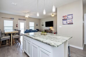 Een moderne keuken met een groot eiland, witte kasten en hanglampen. Er is een zithoek met een houten tafel en stoelen, en een woonkamer met een blauwe bank.