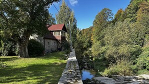 A stone pathway leading to a rustic house with a red roof, surrounded by lush greenery and a small stream. - Izeria 112 Riverside House (Piechowice)