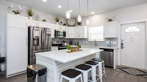 A modern kitchen with white cabinetry, stainless steel appliances, a central island with bar stools, pendant lighting, and decorative plants.
