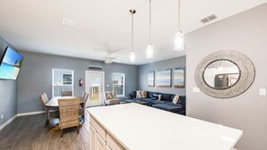 A modern kitchen with a large island, a dining area with wicker furniture, and a living area with a sofa and a TV. - Coastal Escape | Resort Amenities, Pools & Pickleball (Corpus Christi)