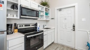 A modern kitchen with white cabinetry, a black stove, a microwave, and a coffee maker. There is a plant on the shelf and a door with a lock.