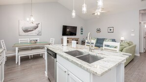A modern kitchen with a marble countertop, white cabinetry, and a built-in dishwasher. There is a dining area with a white table and chairs, and a green sofa in the living room.