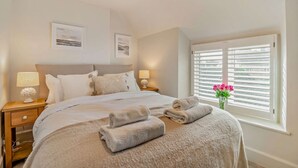 Une chambre à coucher avec un lit, deux tables de chevet avec des lampes, une fenêtre avec des stores et un vase rempli de fleurs.