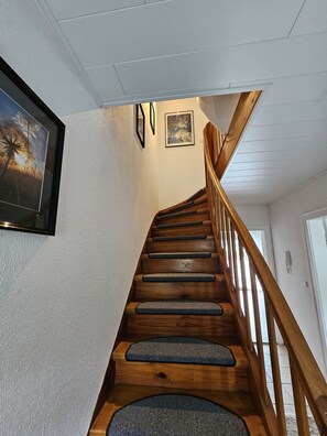 Una escalera de madera con escalones tejidos y barandilla de madera, que lleva a un rellano con un cuadro enmarco y una puerta.