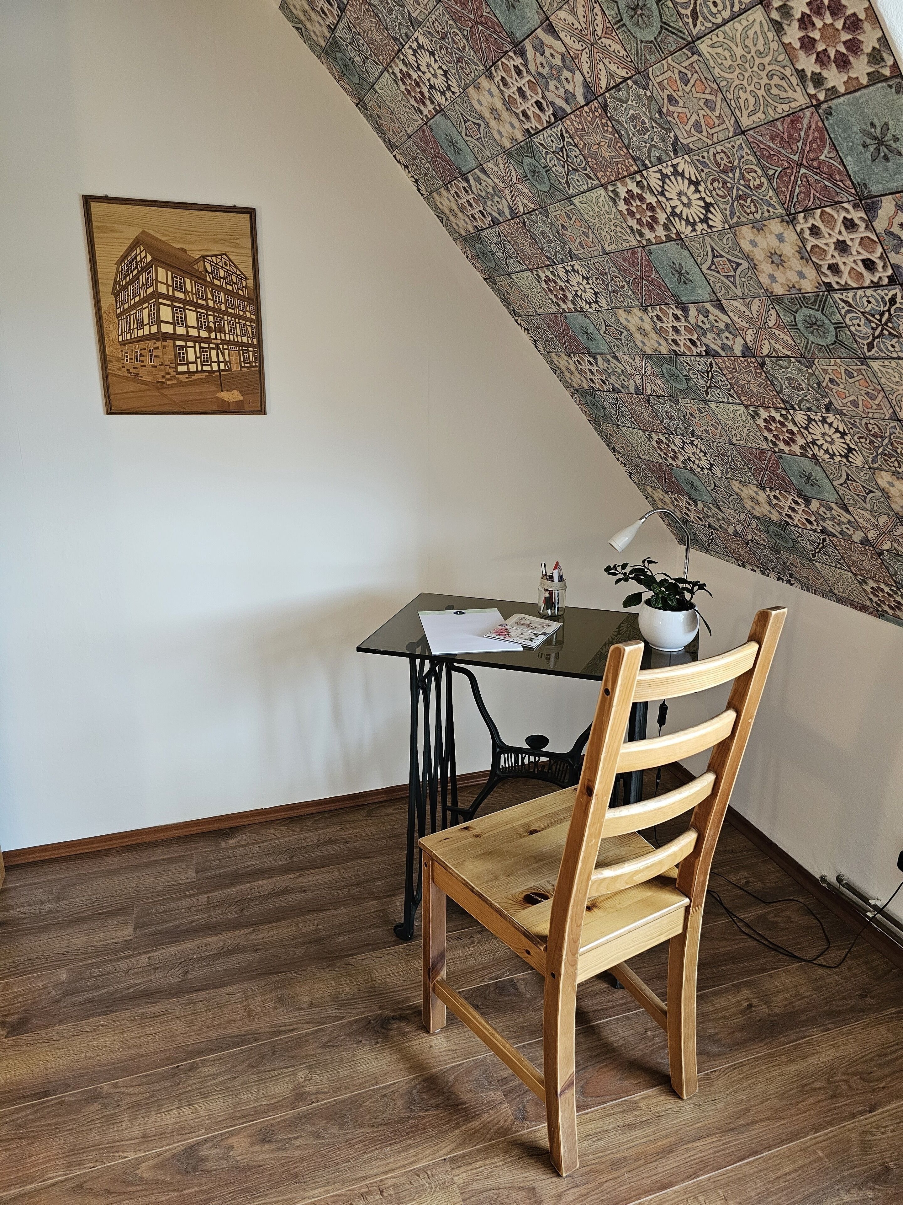 A room with a sloped ceiling, a wooden floor, a table with papers, a chair, and a potted plant.