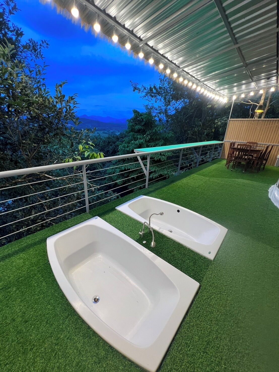 An outdoor area with two white tubs on artificial grass, a wooden table and chairs, and a railing overlooking a natural landscape.