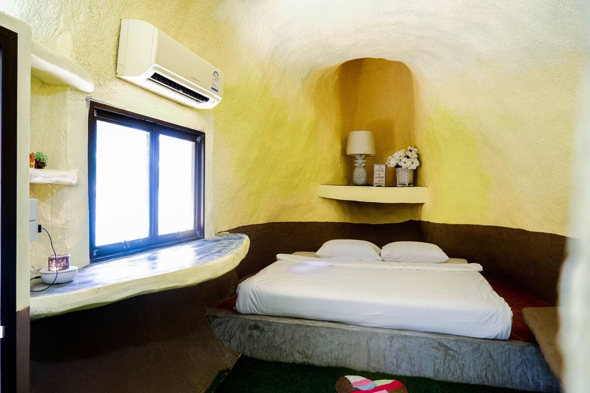 A bedroom with a bed, a window, an air conditioner, a shelf with decorative items, and a small table with a cup and a plant.