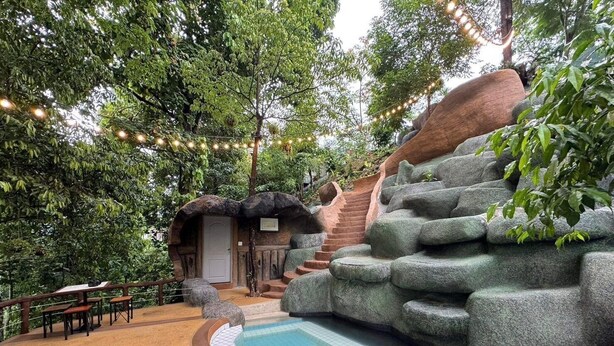 A pool area with a stone waterfall, wooden benches, and a staircase leading up to a thatched-roof structure.