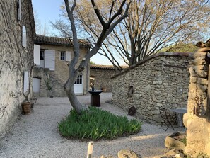 Une cour bordée de murs en pierre, avec un arbre et une petite table entourée de chaises.