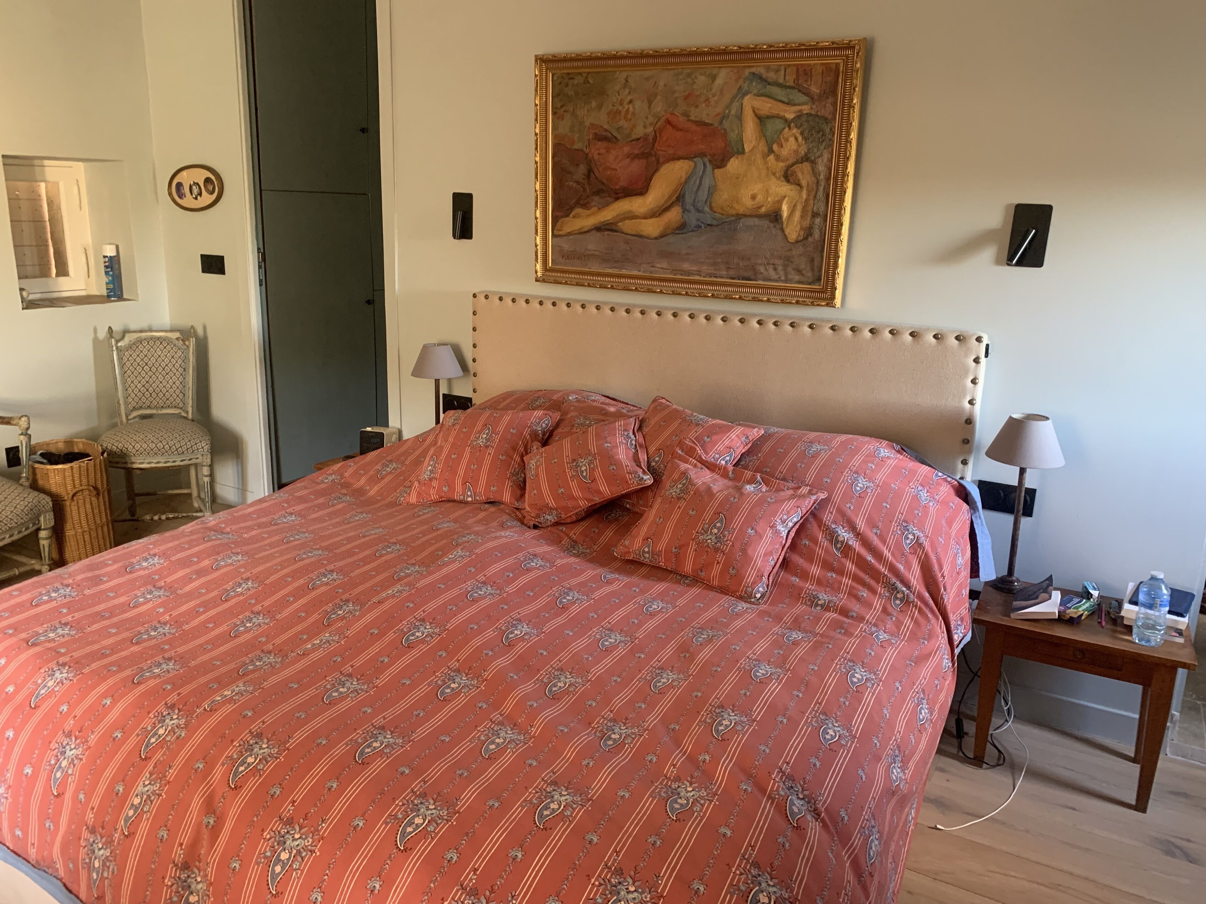 A bedroom with a large bed, a painting on the wall, a chair, a nightstand, and a lamp.