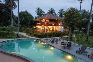 A two-story building with a thatched roof, surrounded by a pool and outdoor seating.
