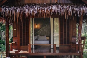 A wooden cabin with a thatched roof, a bed with pillows, and a bench outside.