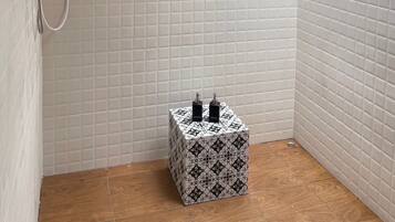 Une petite salle de douche carrelée, équipée d’une pomme de douche et d’un pommeau de douche amovible. Un petit tabouret à motifs se trouve dans un coin.