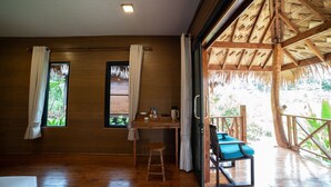 Ein Zimmer mit Holzboden, einem Tisch, zwei mit Vorhängen versehenen Fenstern, einem Stuhl und Blick auf grüne Natur.