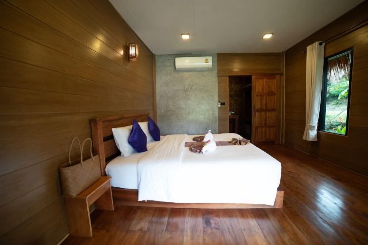 Une chambre avec un plancher en bois, comprenant un lit, des oreillers, une table de chevet et un sac.