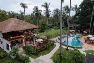 A restaurant with a thatched roof, outdoor seating, and a pool area with lounge chairs and umbrellas.