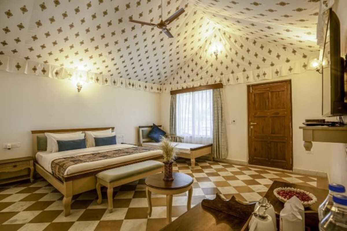 Un dormitorio con suelo a cuadros, una cama con colcha estampada, una mesita de noche con un florero y una puerta de madera.