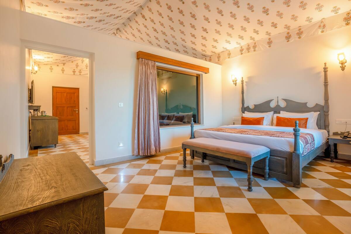 A bedroom with a checkered floor, a wooden bed with white and orange bedding, a bench, a window with curtains, and a door leading to another room.