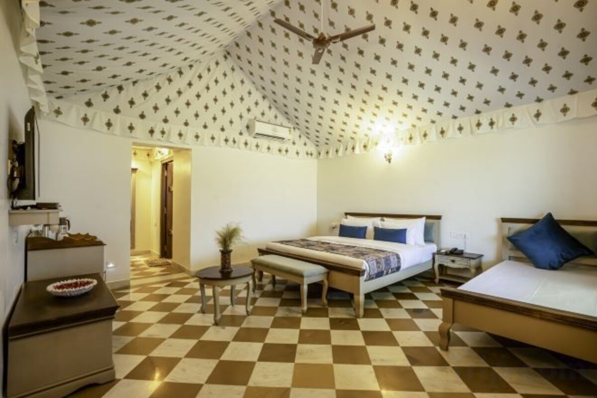 Un dormitorio con piso de cuadros, una cama con almohadas azules, una mesita de noche y un escritorio con una lámpara.