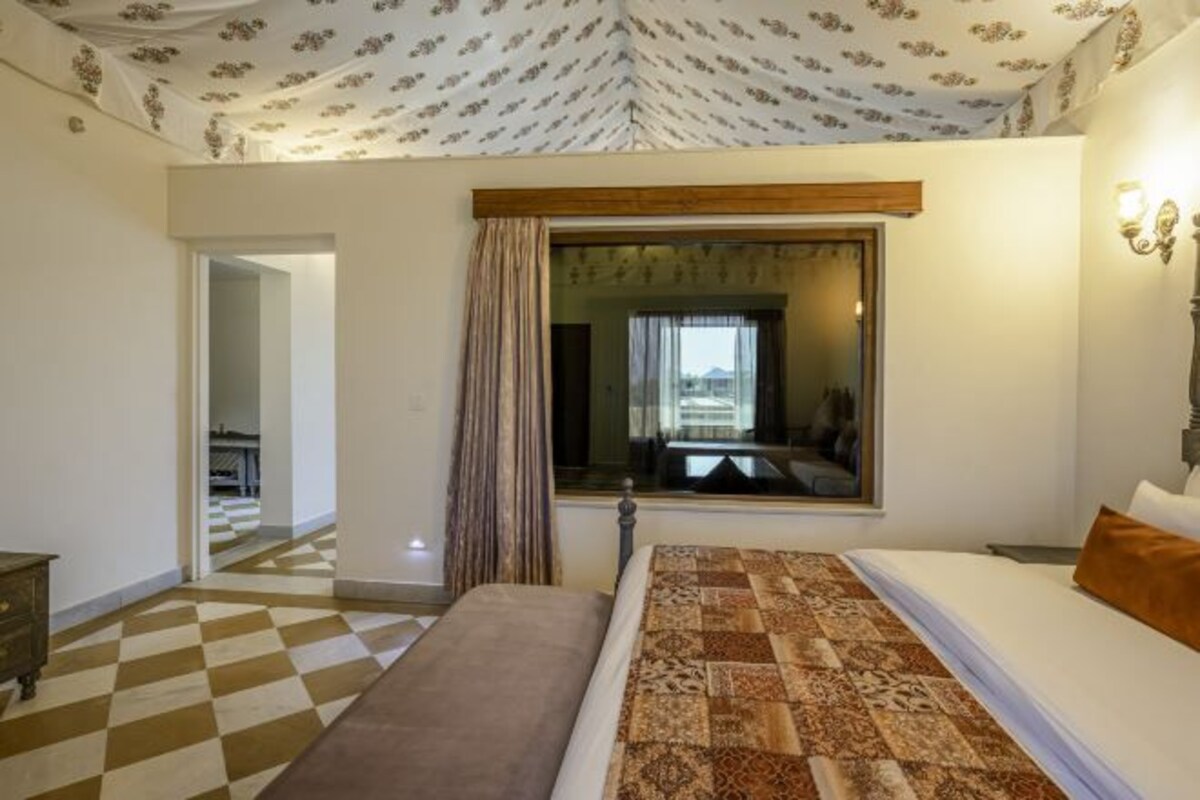 A hotel room with a checkered floor, a bed with a patterned bedspread, a window with curtains, and a view of another room.