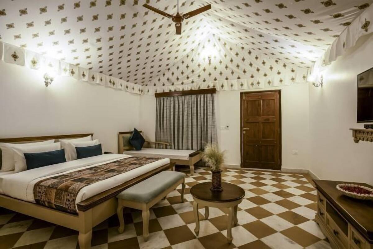 A room with a checkered floor, a bed with a patterned bedspread, a bench, a small table with a vase, a wooden door, and a wall-mounted shelf.