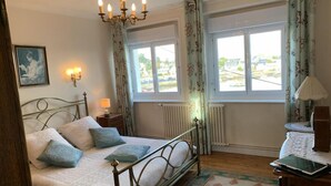 A bedroom with a bed, a chandelier, a window with curtains, a radiator, and a bedside table with a lamp. - Maison Ploubazlanec Pour 6 Personnes (Ploubazlanec)