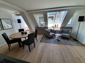 Un salon moderne avec un coin repas, comprenant une table ronde, des chaises, un canapé et une petite table basse. De grandes fenêtres laissent entrer la lumière naturelle et offrent une vue sur l’extérieur.