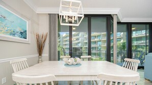 Un coin repas avec une table et des chaises blanches, une pièce maîtresse décorative et une vue sur un immeuble d’habitation à travers de grandes fenêtres.