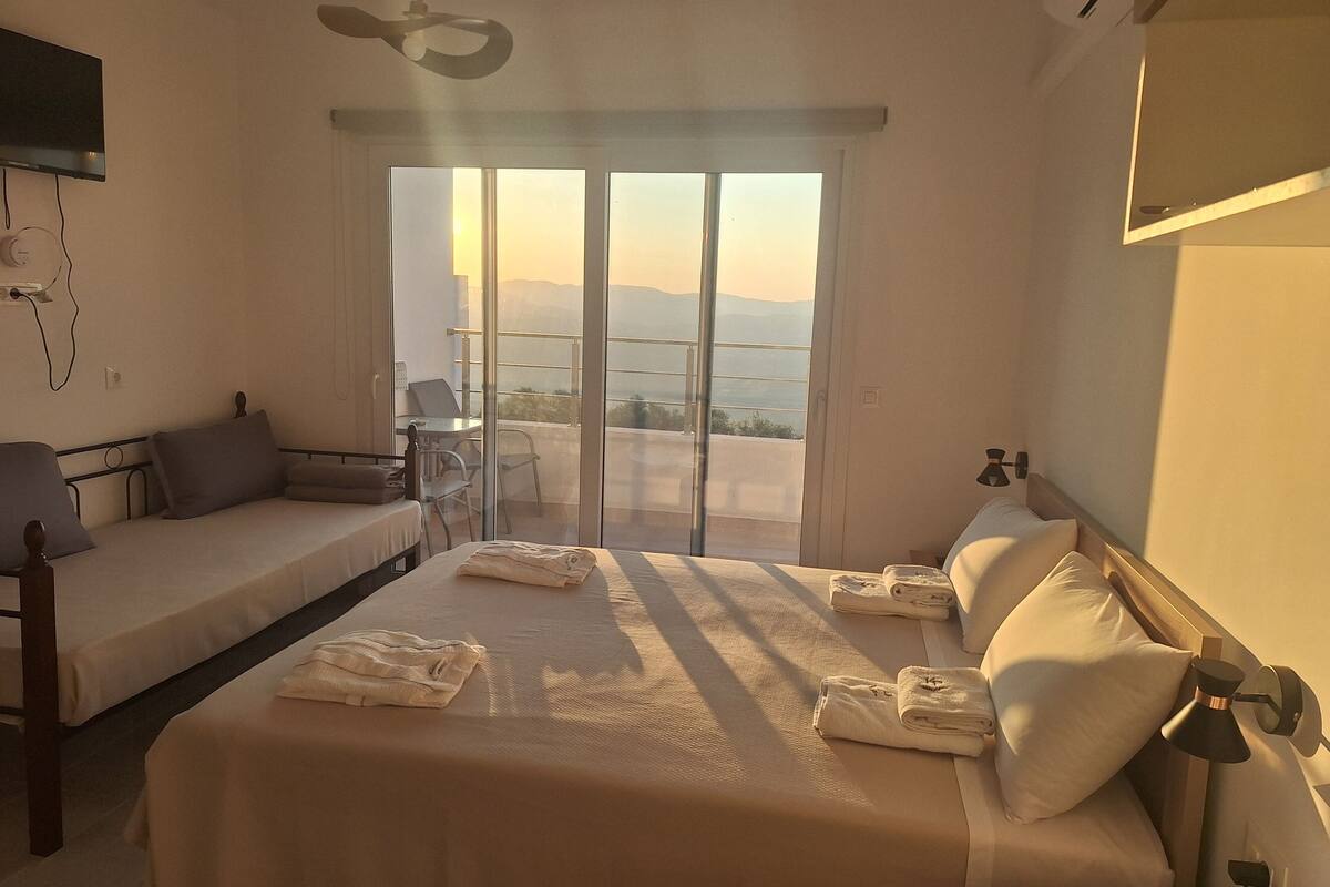 Een hotelkamer met een bed, een slaapbank, een bureau en een grote schuifdeur naar een balkon met uitzicht op de bergen.