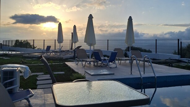 Un espace aménagé au bord de la piscine, avec des chaises longues et des parasols, une table et une vue sur le coucher de soleil sur l’eau.