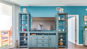 A teal-colored entertainment center with a flat-screen TV, surrounded by shelves with decorative items and a wicker basket. - Palms of Destin Resort Condo w/ Private Balcony! (Destin)
