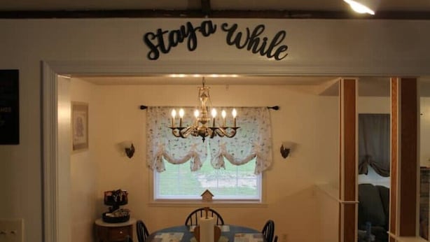 A dining area with a chandelier, a table set for four, and a sign above reading "Stay a While".