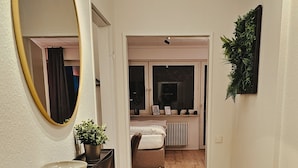 Ein Flur mit Holzboden, einem schwarzen Konsolentisch mit Schale und Topfpflanze, einem großen Spiegel und einer Tür, die in ein Schlafzimmer mit Bett und Heizkörper führt.