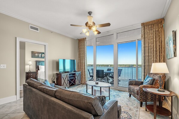 A spacious living room with a ceiling fan, a television, and a balcony with a view of the water.