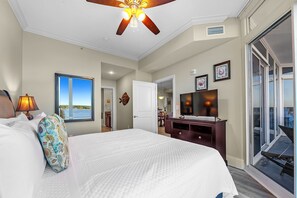 A bedroom with a large bed, a ceiling fan, a TV, and a view of the outdoors.