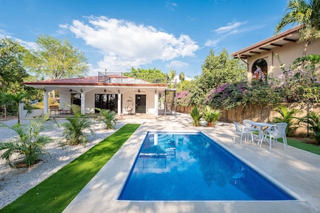 A backyard with a swimming pool, a covered patio, and a garden with various plants and trees.
