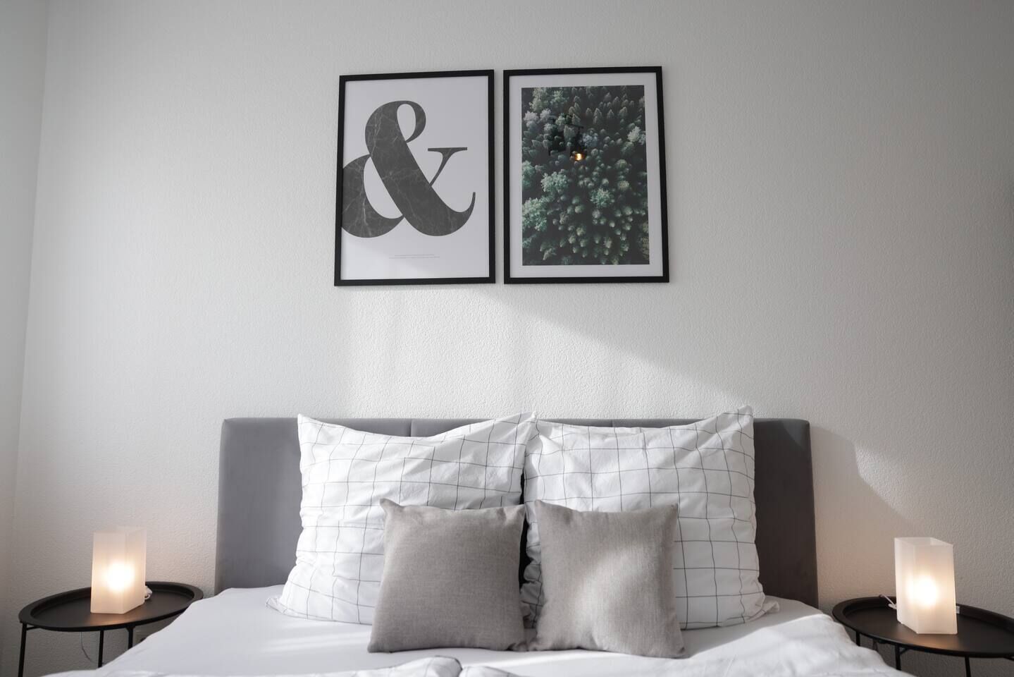 Een keurig opgemaakt bed met twee kussens, een nachtkastje met een lamp, en twee ingelijste kunstwerken aan de muur.