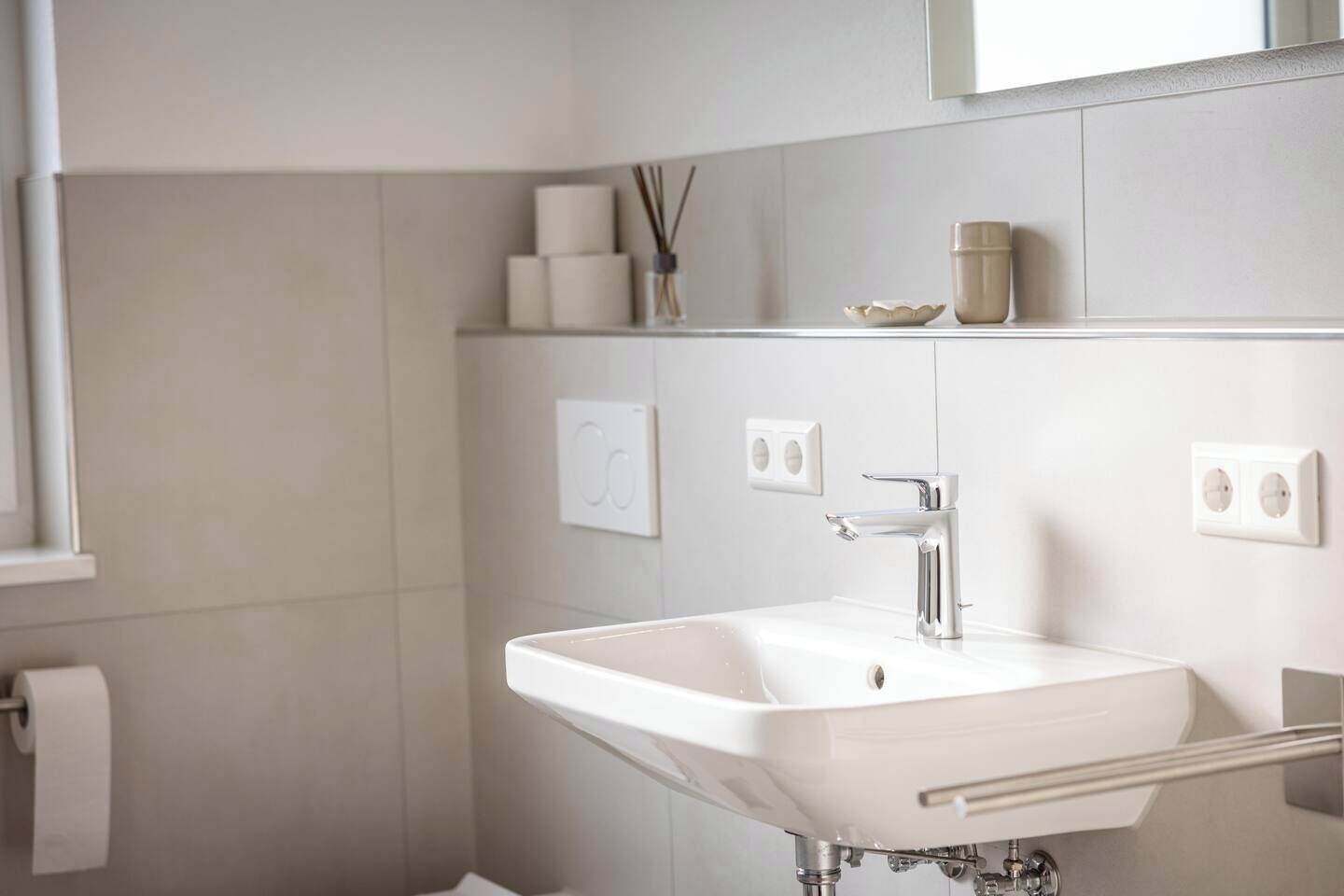 Ein modernes Badezimmer mit einem weißen Waschbecken, einem Chromarmaturen und einem Handtuchhalter. Zwei Wandleuchten schalter und ein kleines Regal mit Dekorationen sind vorhanden.