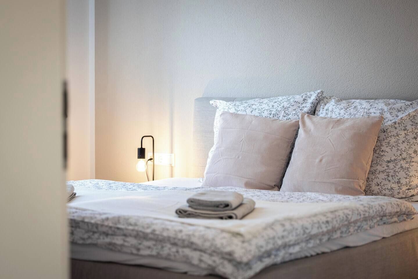 A neatly made bed with a white duvet, two pillows, and a folded towel on the bed. There is a wall-mounted light fixture to the left of the bed.